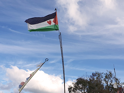 An image of a hand sewn Palestinian flag flying on a flagpole welded by Graham Hughes. The pole is welded from recycled old rebar. Flying flags in the top paddock as simply as possible is a design challenge. It is very windy up there! The flag is a statement about Graham's solidarity with the people of Palestine, and while in the face of many seeming to tolerate the genocide and historical injustice, he does not. This is not shared by others, and that's okay. The heart sewn onto the flag expresses that.